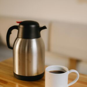 A stainless steel thermos and coffee mug filled with black coffee on a wooden table indoors.