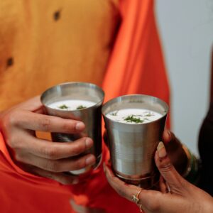 Close-up of two hands holding traditional Indian buttermilk in metal glasses, indoor setting.